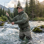 Man fly fishing in a river surrounded by trees and mountains, wearing a green jacket and pants.